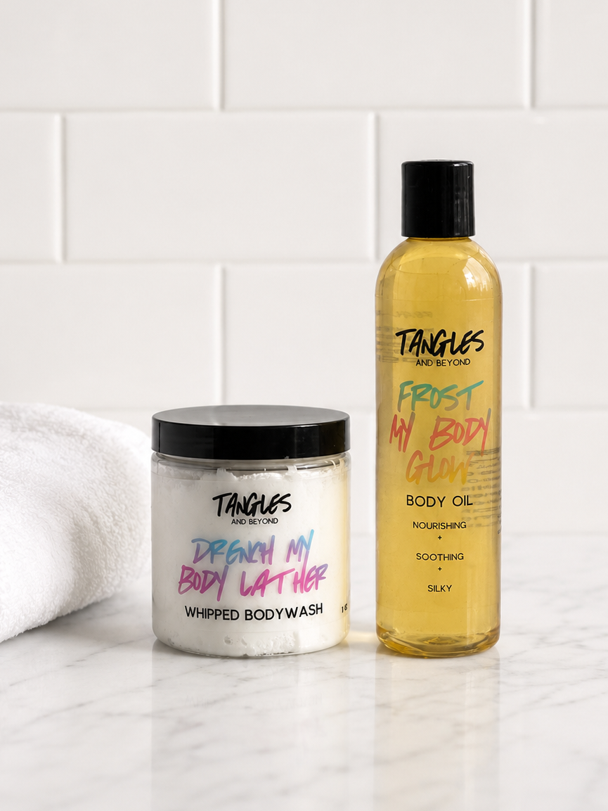 Two Tangles and Beyond  Body Care products on a white marble surface with a white subway tile background: a jar of Dreamy Body Butter Whipped Body Wash and a bottle of Frosty Body Oil Body Oil, with a folded white towel on the left.