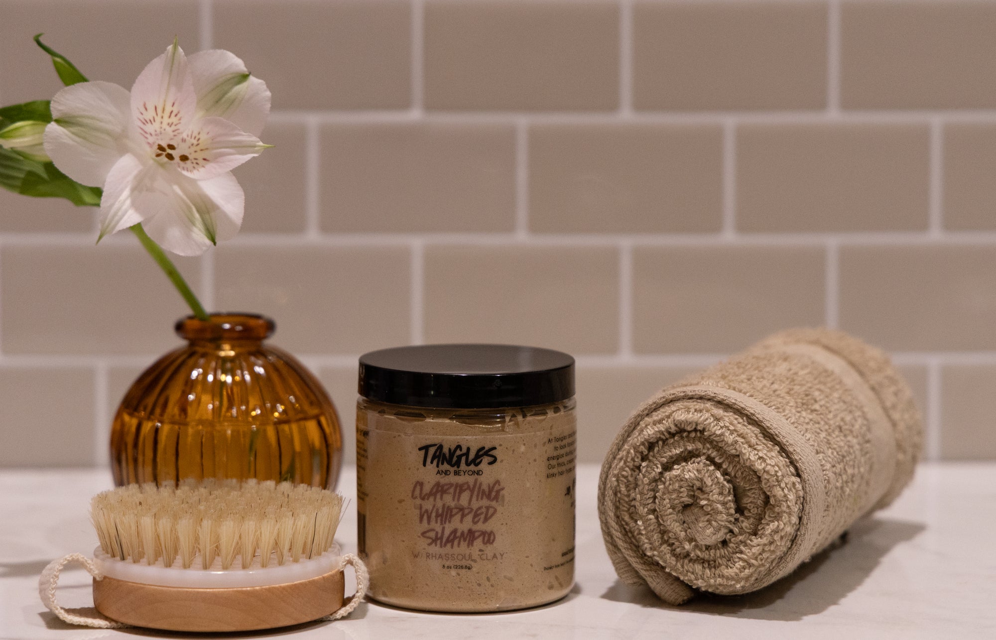 Cosmetic products including a jar, scrubber, and rolled towel on a surface with a tiled wall background.