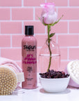 Bottle of Tangles hair product with a pink rose, scrubber, and bowl on a pink tiled background