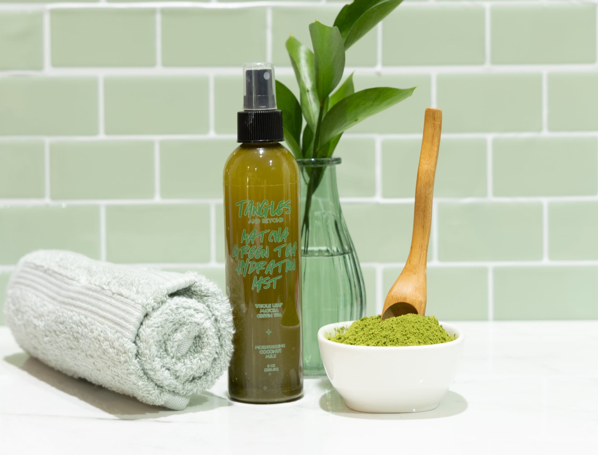 Bottle of green hair product with towel, bowl of green powder, and wooden spoon on a light green tiled wall background.